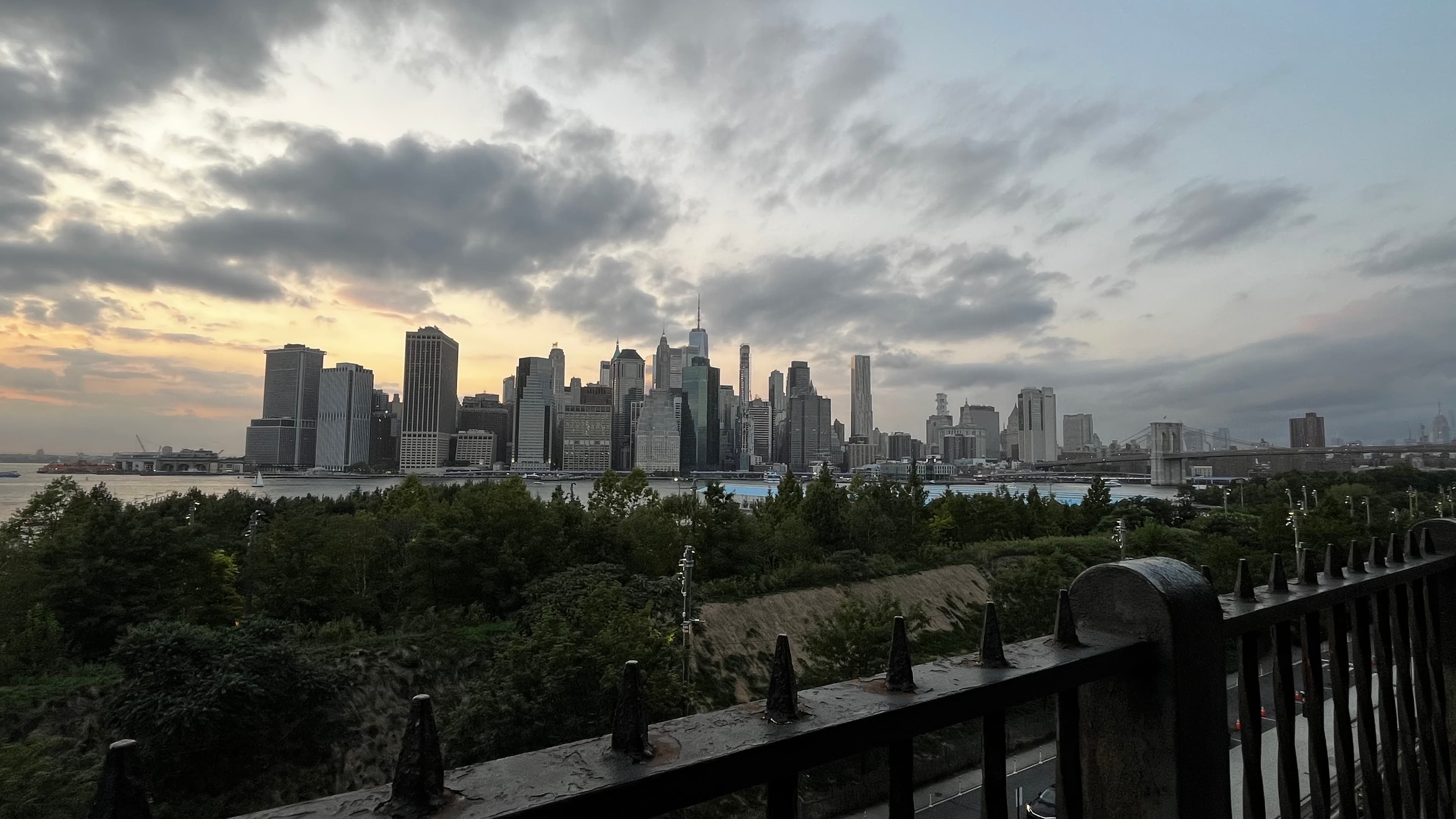 Lower Manhattan skyline at sunset under dramatic clouds, framed by trees and a spiked railing.